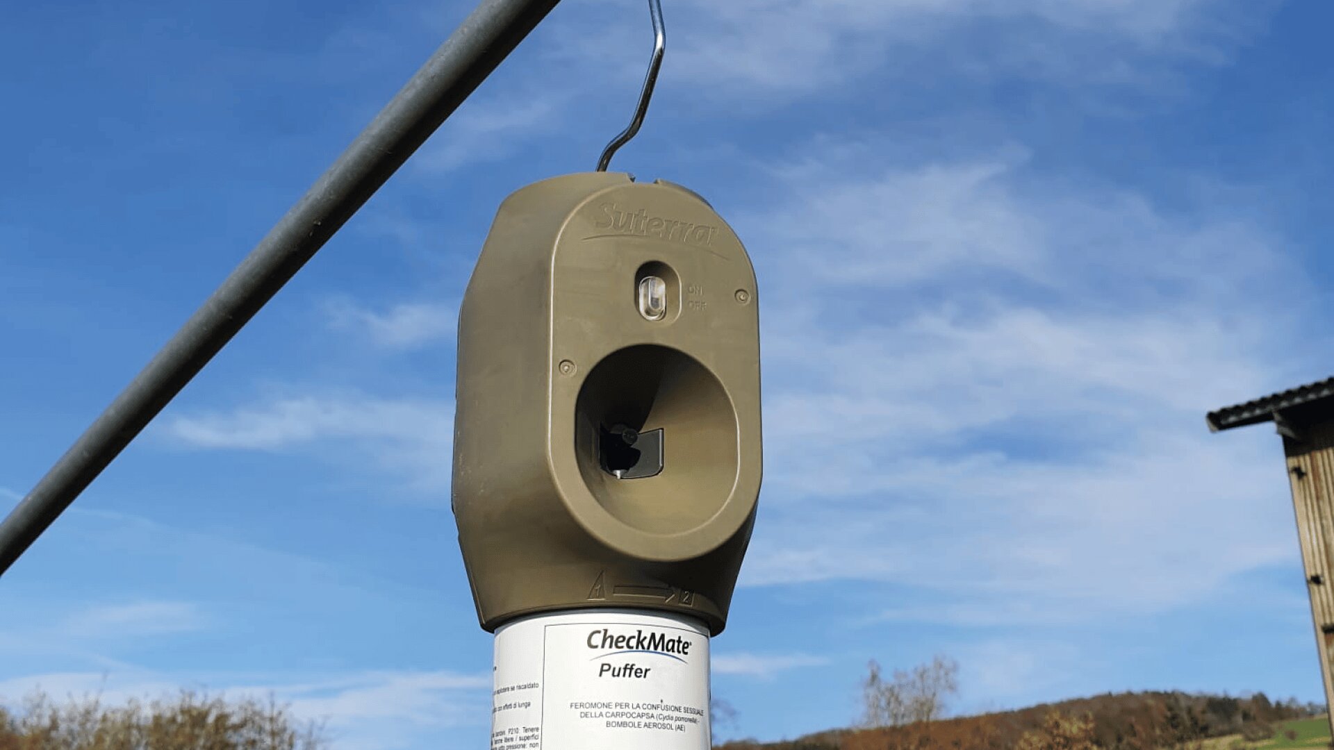 Nahaufnahme eines bräunlichen Insektenpheromonspenders mit der Aufschrift CheckMate Puffer, der im Freien an einer Metallstange hängt, mit blauem Himmel und Bäumen im Hintergrund.